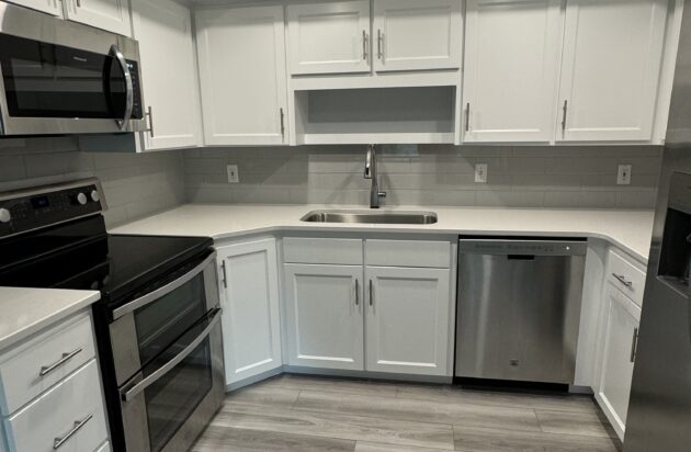 A modern kitchen with gallery-like elegance features white cabinets, stainless steel appliances, and a double sink. The recessed lighting in the ceiling highlights the space, while a light wood floor adds warmth and style.